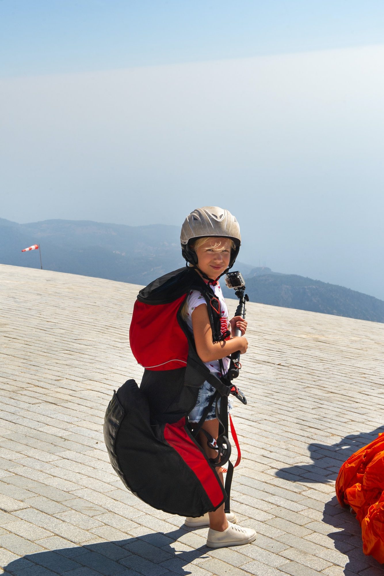 a little girl in paragliding gear and with a camera stands on the edge of mount babadag turkey e1655171742105